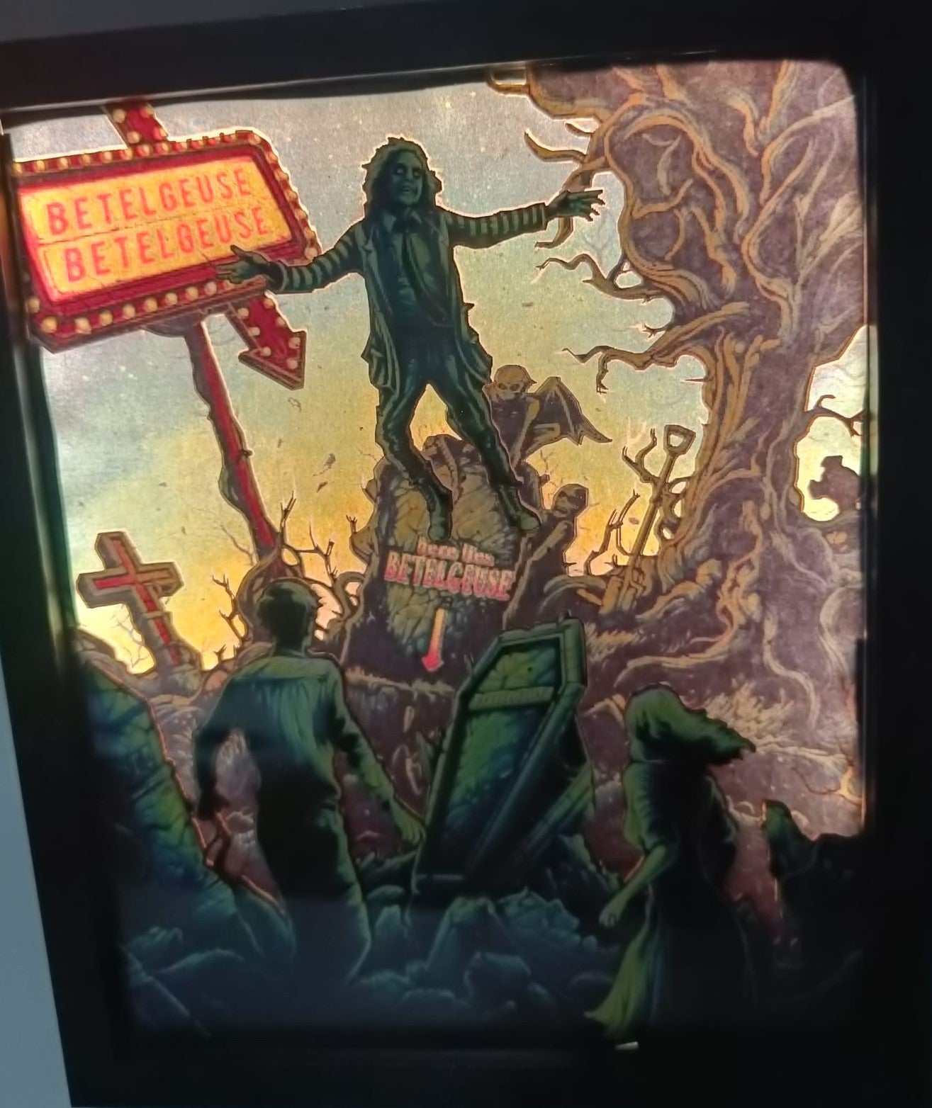 Un'illustrazione stilizzata e inquietante di L'Atelier di Zelda mostra una figura su un tumulo etichettato Beetlejuice dal film. Alberi contorti, lapidi e un cartello rosso al neon Betelgeuse creano la scena del bio-esorcista con figure in silhouette e una bara che evocano lo stile di Tim Burton.
