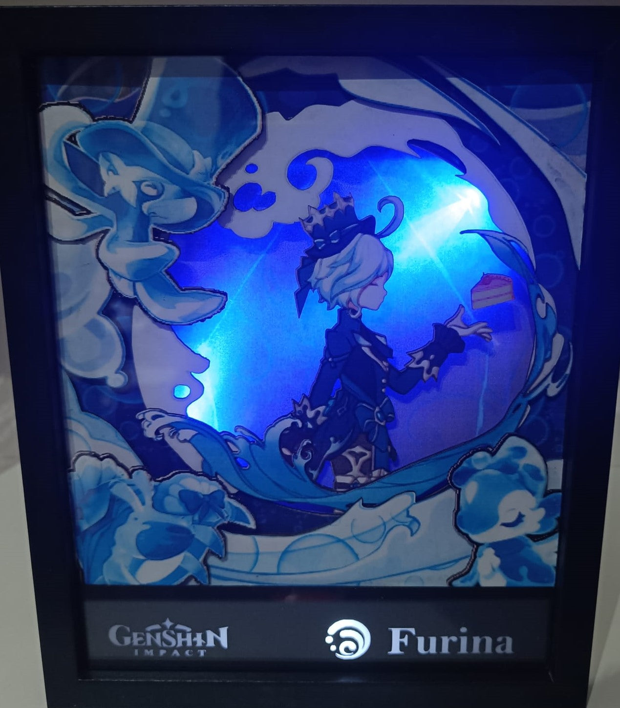 Il pezzo di L'Atelier di Zelda, "Genshin Impact - Furina", presenta una foto incorniciata con una radiosa luce blu. Furina tiene elegantemente un piccolo oggetto tra gli elementi d'acqua di Fontaine, con una silhouette di cappello e il logo del gioco sullo sfondo.