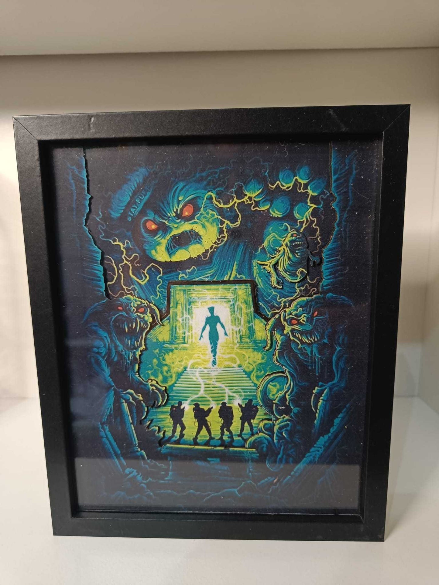 Un'illustrazione incorniciata realizzata da L'Atelier di Zelda per il film "Ghostbuster" mostra quattro figure simili ad acchiappafantasmi di fronte a una porta luminosa da cui emerge una persona, circondata da volti mostruosi e creature con occhi rossi e viticci, che evocano il regno di Gozer.