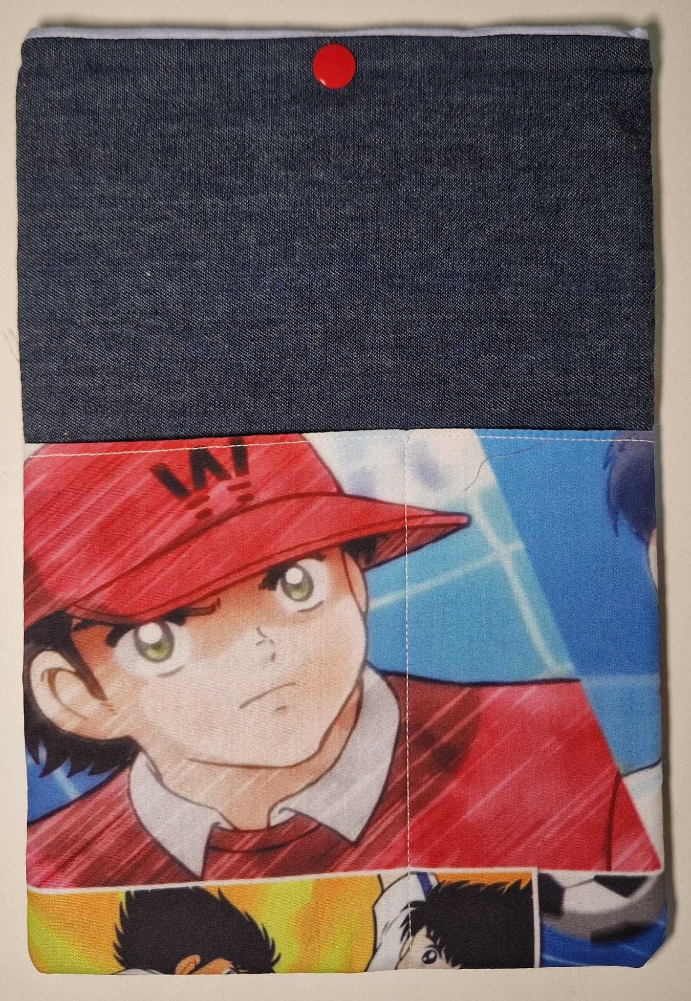 La custodia in tessuto, chiamata "Holly e Benji - Benji (Portalibro)" di L'Atelier di Zelda, raffigura un personaggio anime dagli occhi spalancati di "Captain Tsubasa" con un berretto rosso. Il suo risvolto superiore in stile denim e i colori vivaci catturano lo spirito energico di questa iconica storia a tema calcistico.