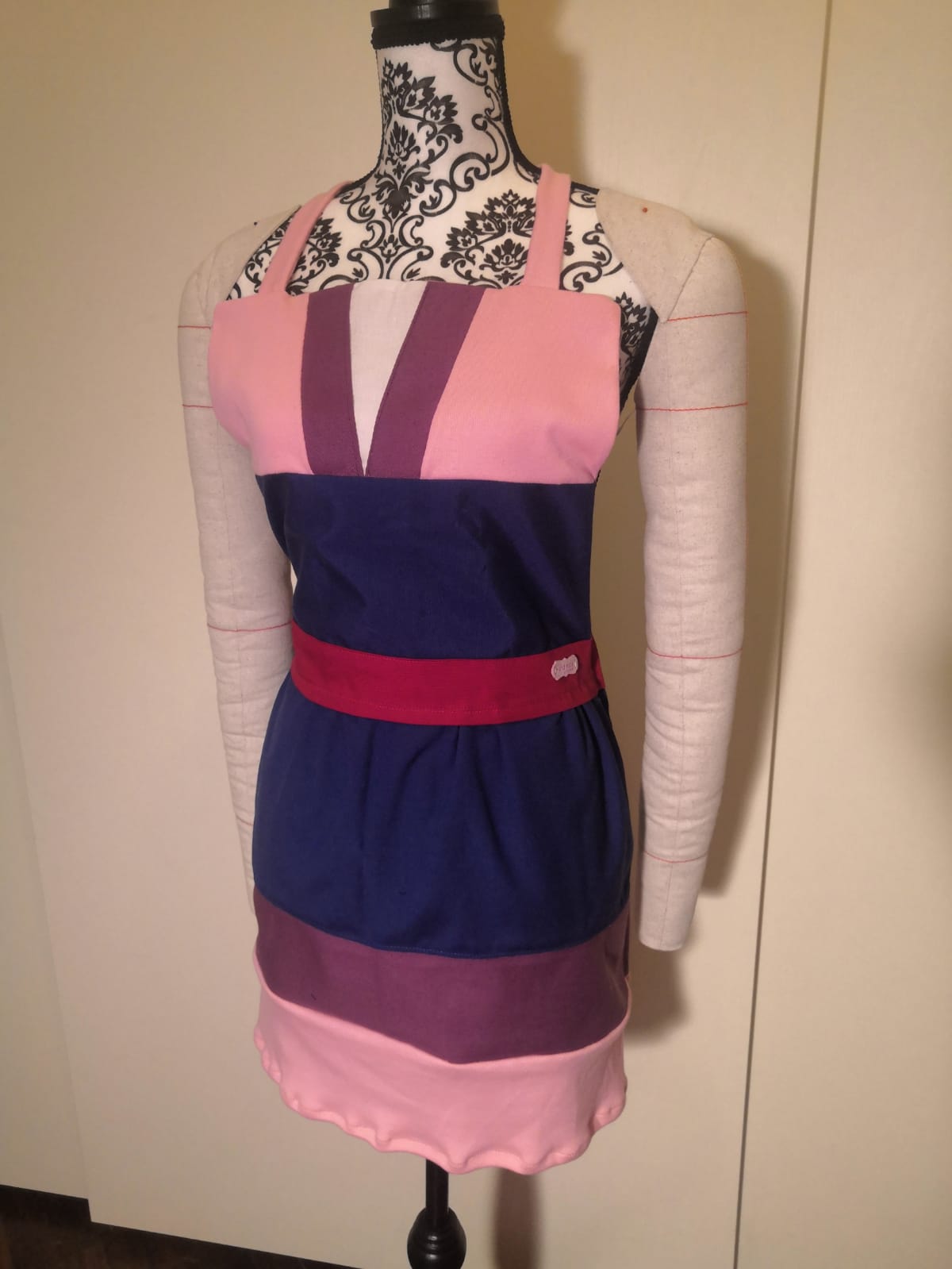 Un manichino presenta il "Grembiule fatto a mano ispirato a Mulan" di L'Atelier di Zelda, caratterizzato da vivaci strisce orizzontali rosa, blu e viola con un'elegante scollatura rosa e una cintura color porpora, su uno sfondo di un motivo floreale nero.